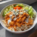 A close-up of a delicious buffalo chicken bowl featuring shredded orange buffalo chicken over brown rice, topped with blue cheese crumbles and ranch dressing.