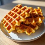 A close-up stack of golden brown, freshly made high protein waffles resting on a light-colored plate.