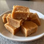 A close-up stack of rich, light brown squares of homemade penuche fudge resting on a white plate.