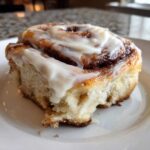 A close-up of a fluffy, freshly baked cinnamon roll generously topped with white cream cheese icing.