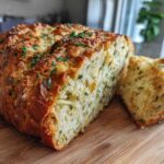 A freshly baked loaf of Italian herb and cheese bread, sliced to show the cheesy interior, resting on a wooden cutting board.