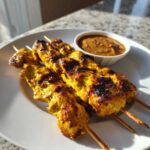 Four golden, grilled chicken satay skewers served on a white plate next to a small bowl of peanut dipping sauce.