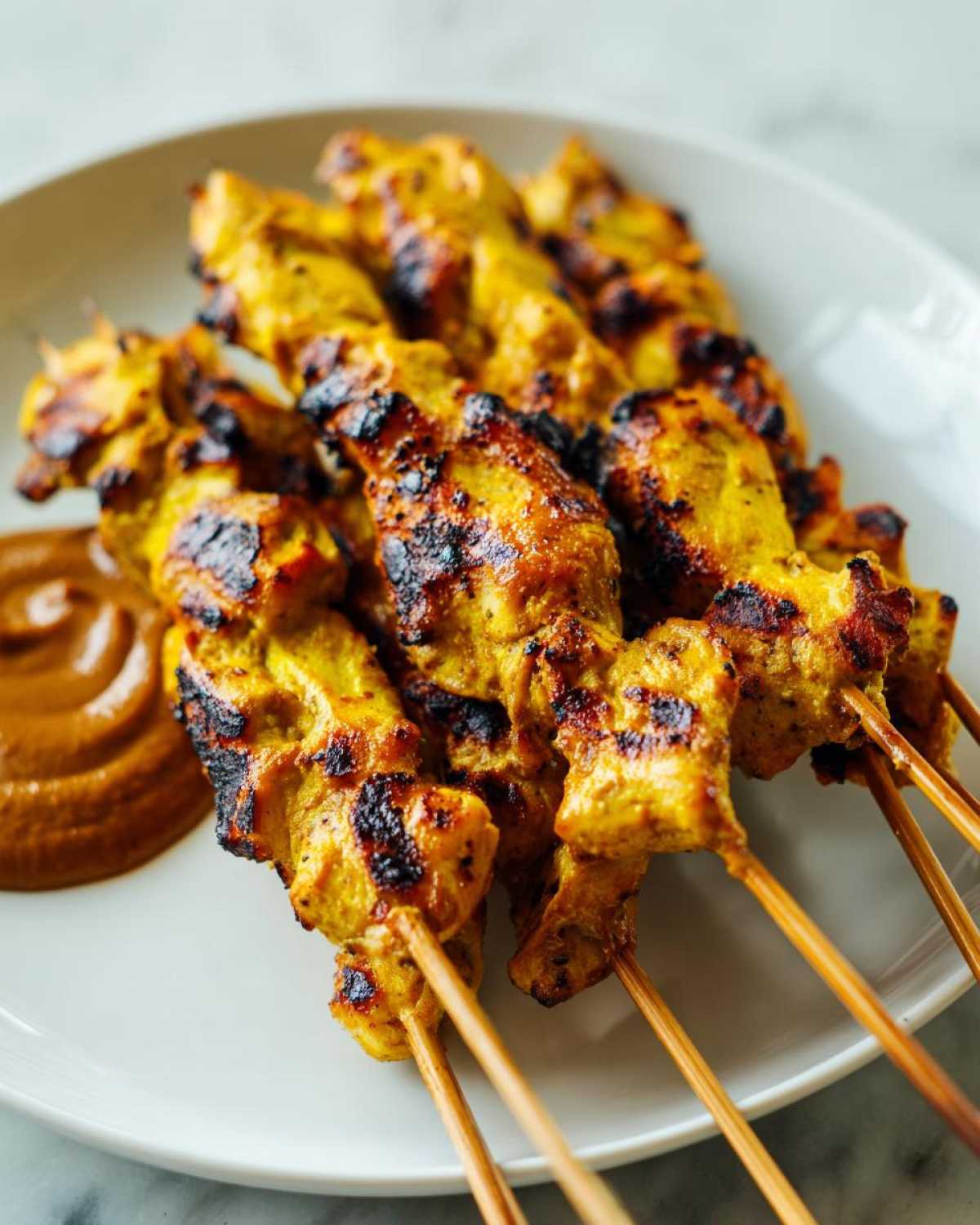 Close-up of grilled, yellow-marinated chicken satay skewers served with a side of peanut dipping sauce.