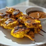 Close-up of grilled, yellow-marinated chicken satay skewers served with a side of rich peanut dipping sauce.