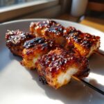 Close-up of juicy chicken skewers coated in a dark, sticky, chili-flecked glaze resting on a white plate.