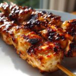 Close-up of three skewers featuring juicy, glazed, and slightly charred chicken skewers resting on a white plate.
