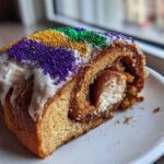 A close-up of a slice of cream cheese filled king cake, topped with white icing and purple, gold, and green Mardi Gras sprinkles.