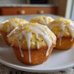 Four golden lemon poppy seed muffins topped with white glaze and lemon zest, resting on a white plate.