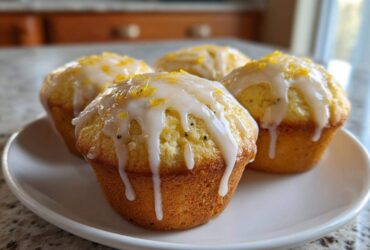 Four golden lemon poppy seed muffins topped with white glaze and lemon zest, resting on a white plate.