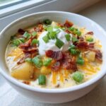 A creamy bowl of loaded baked potato soup topped with sour cream, bacon bits, shredded cheese, and green onions.