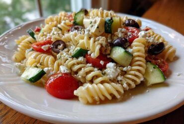 A serving of vibrant Mediterranean pasta salad featuring rotini pasta, cherry tomatoes, cucumber, olives, and feta cheese.