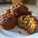 A close-up of several moist bran muffins on a white plate, one cut open showing the texture.
