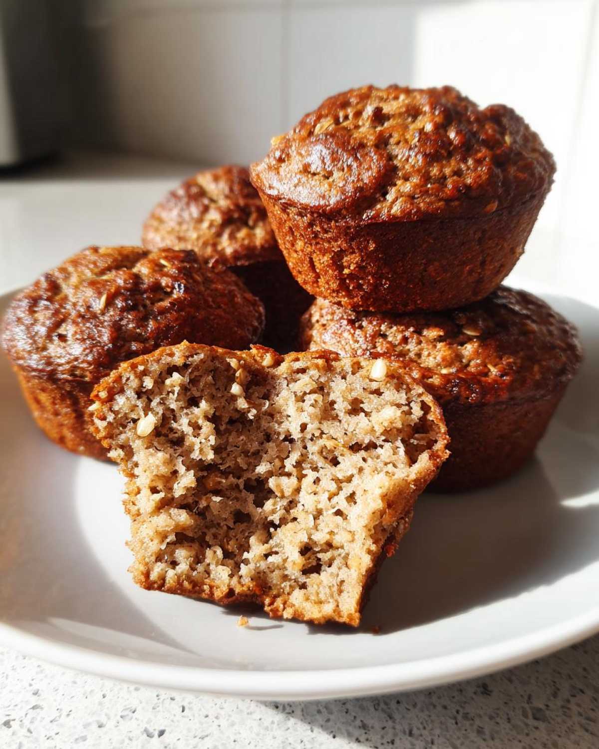 Close-up of several moist bran muffins stacked on a plate, one cut open showing the textured, light brown interior.