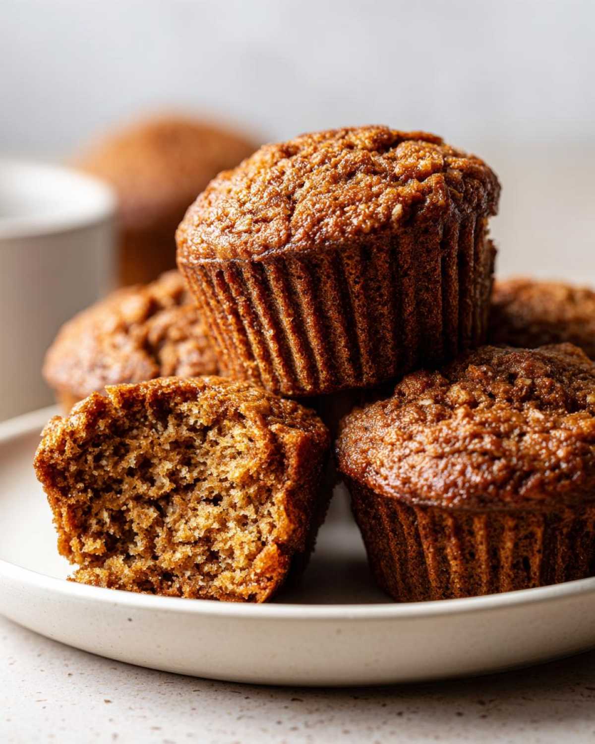 Close-up of several moist bran muffins stacked on a white plate, one muffin is broken open showing the texture.