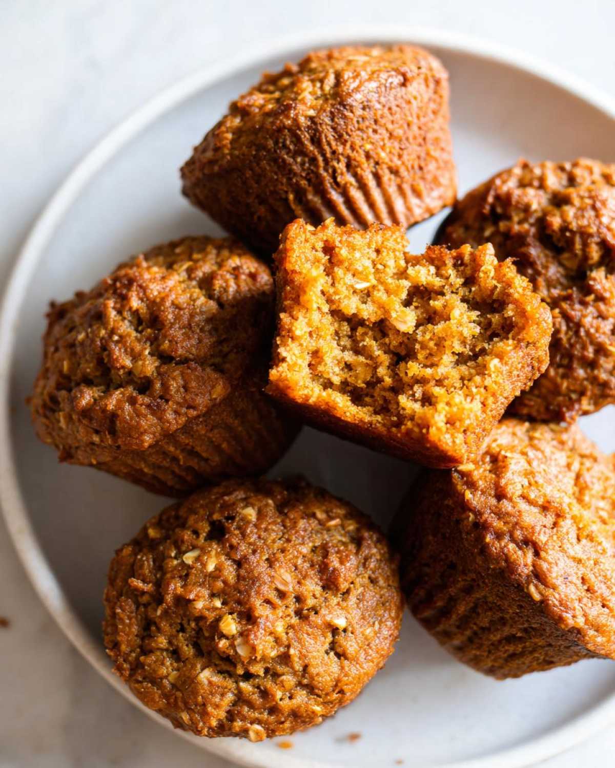 Close-up of several moist bran muffins piled on a plate, one is broken open showing the texture.