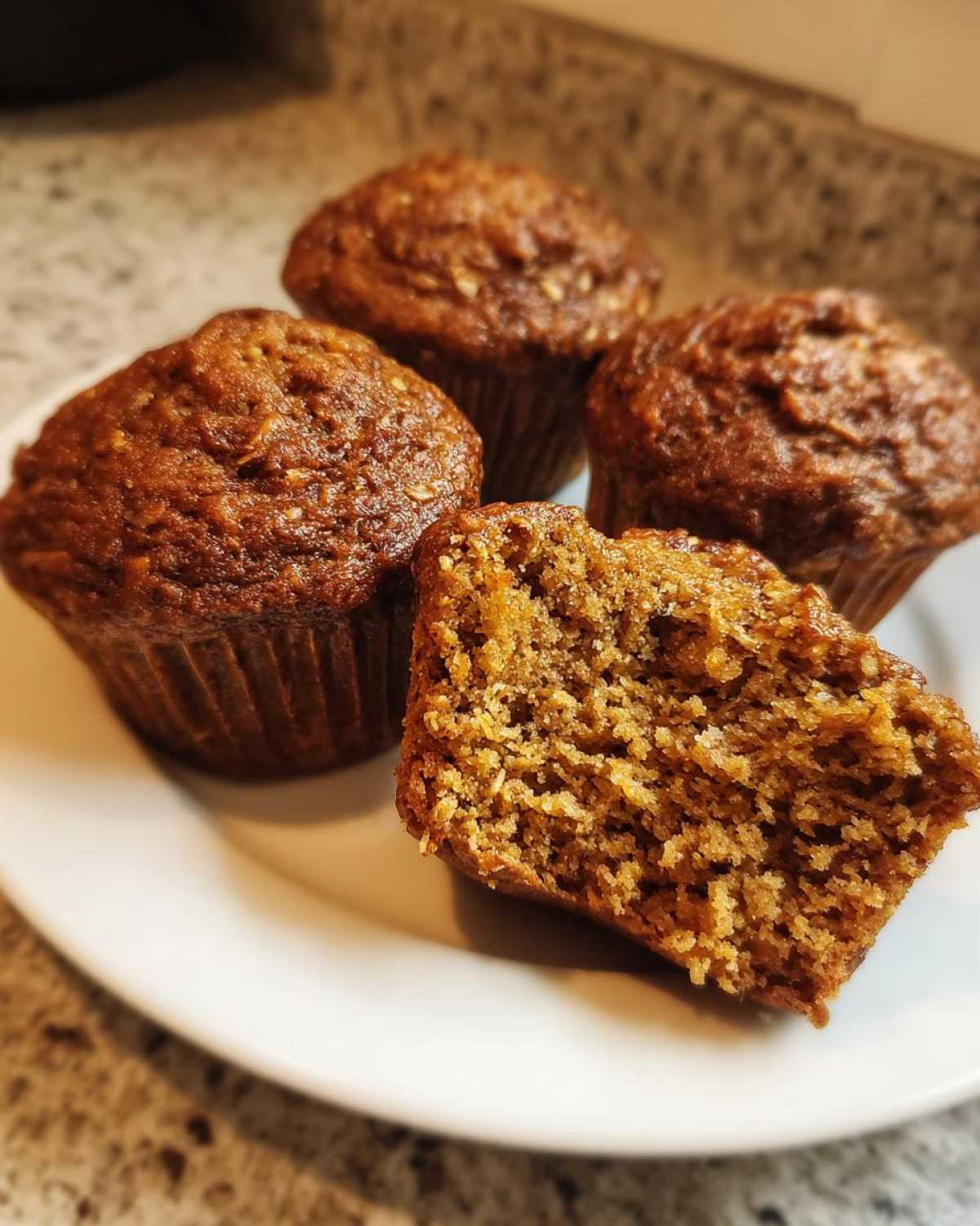 Four freshly baked bran muffins on a white plate, one split open showing the moist, textured interior.