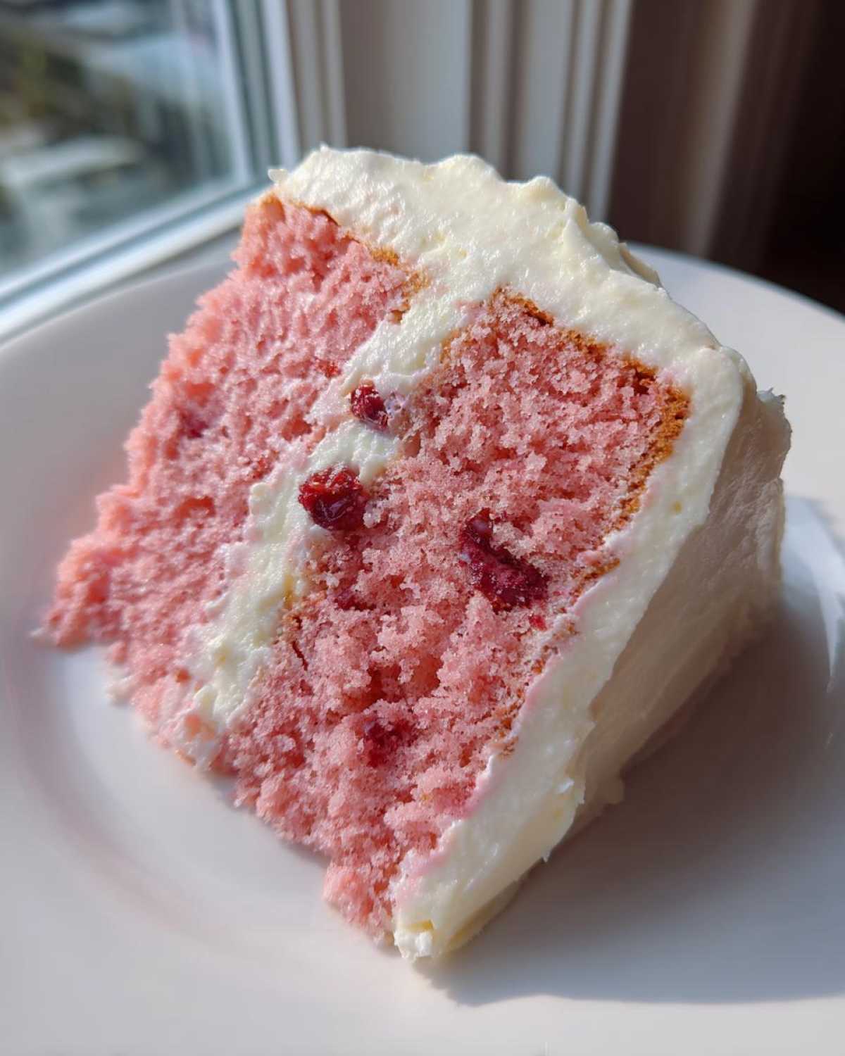 Close-up of a slice of moist pink cherry cake layered with white frosting and visible cherry pieces.