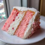 A close-up of a moist slice of pink Cherry cake layered with white frosting and visible cherry pieces.