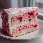 A generous slice of moist cherry cake featuring layers of white cake studded with bright red cherries and layered with white and pink frosting.
