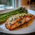 A perfectly cooked salmon recipe fillet topped with garlic and parsley, served alongside roasted asparagus on a white plate.