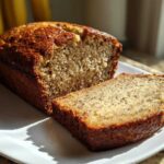A loaf of moist greek yogurt banana bread with one thick slice cut and resting beside it on a white plate.