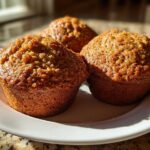 Close-up of three golden brown, moist honey bran muffins sitting on a white plate in bright sunlight.