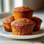 Four perfectly baked, dark brown honey bran muffins stacked on a white plate, showing their moist texture.