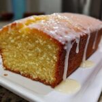 A close-up of a moist lemon pound cake loaf topped with thick white glaze dripping down the sides onto a white plate.