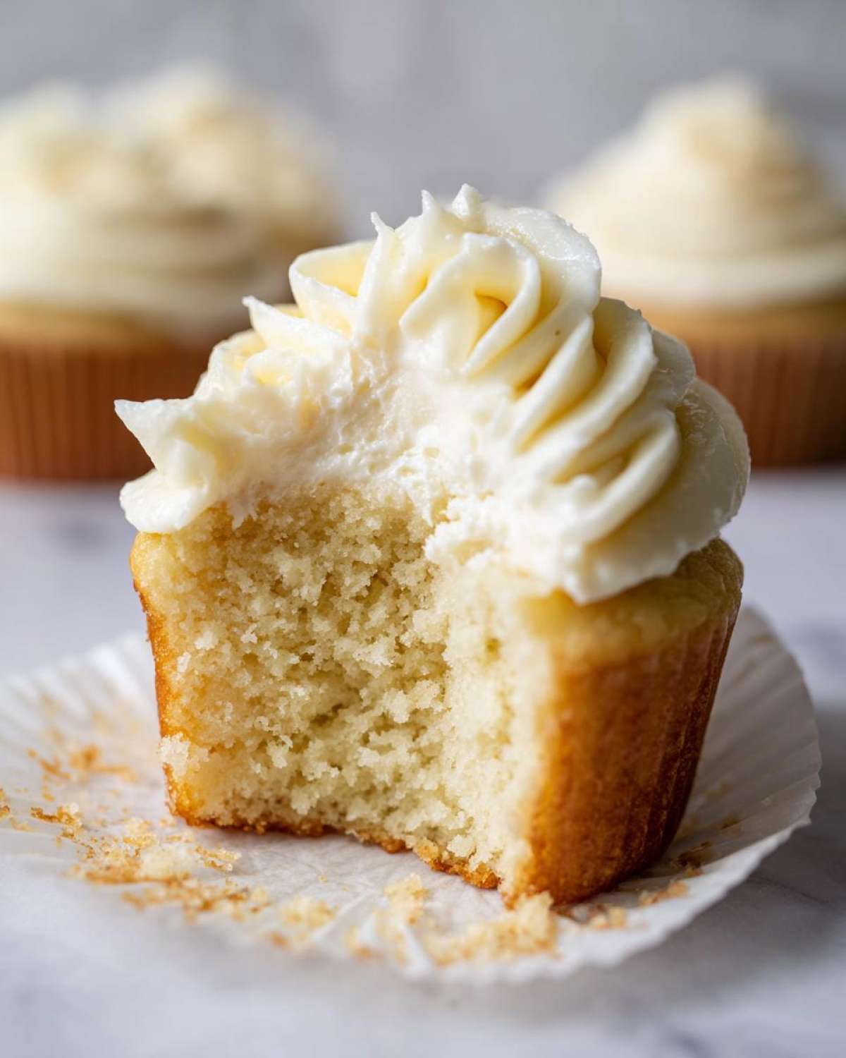 Close-up of a vanilla cupcake with a bite taken out, showing the moist crumb and swirl of white vanilla frosting.