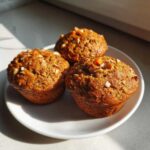 Three freshly baked morning glory muffins topped with chopped nuts and carrot pieces, sitting on a white plate.
