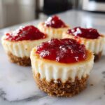 Four individual no-bake cheesecake bites with graham cracker crusts topped with bright red strawberry jam.