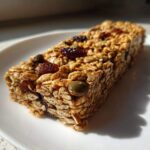 A single, rectangular no bake granola bars made with oats, visible dried cranberries, and pumpkin seeds, sitting on a white plate.