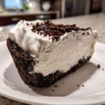 A close-up of a rich slice of no-bake ice cream pie featuring a dark crust, creamy filling, and whipped topping sprinkled with crumbs.