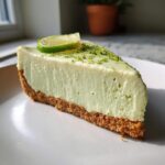 A close-up slice of creamy, pale green no-bake key lime cheesecake on a white plate, topped with lime zest and a lime slice.