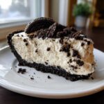 A thick slice of creamy no-bake Oreo cheesecake with a dark chocolate crust, topped with crumbs and a whole Oreo cookie.
