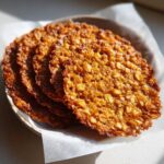 Close-up of several thin, crispy oatmeal lace cookies stacked on a plate with parchment paper.