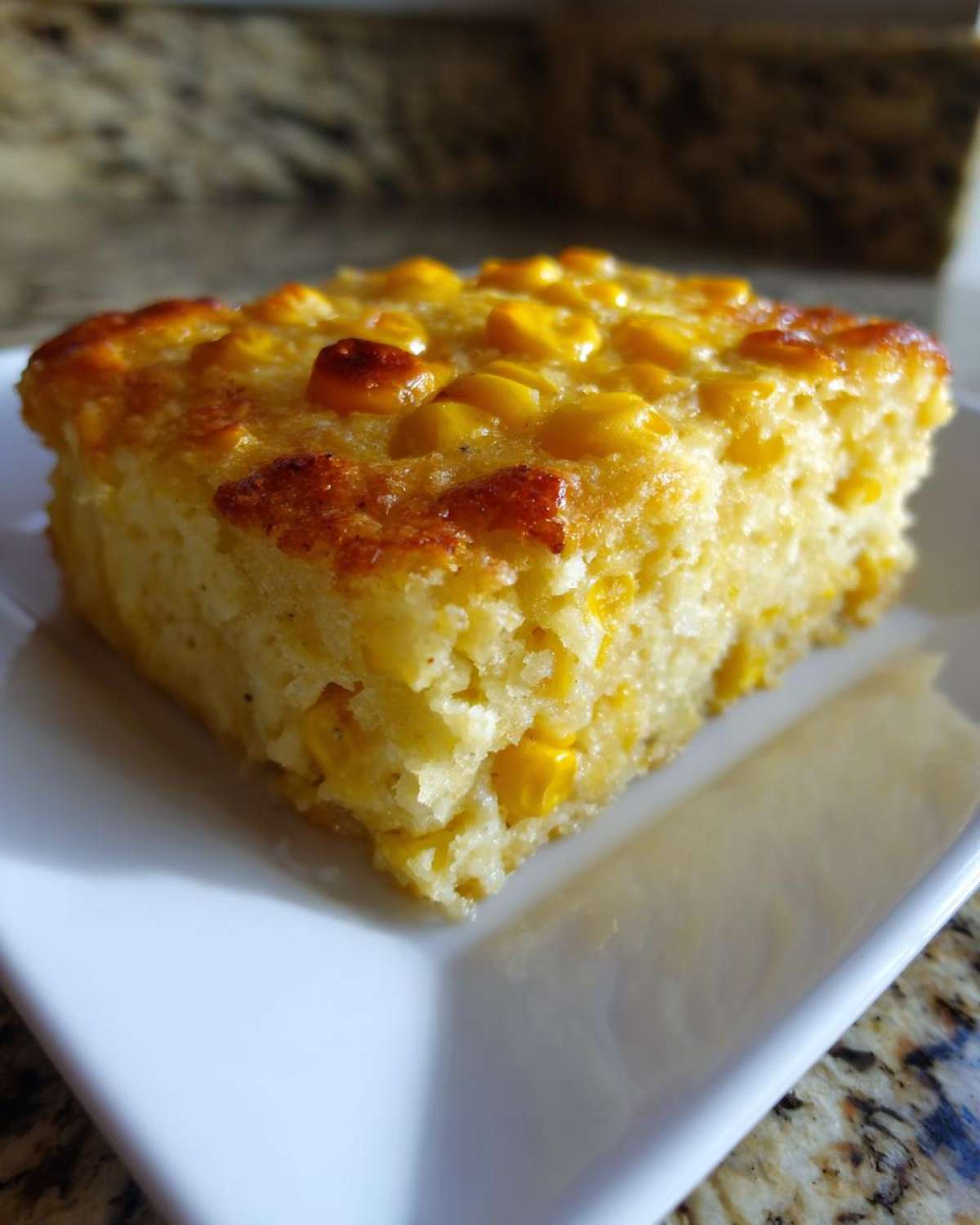 A close-up of a square slice of moist, golden Amazing 1-Bowl Corn Casserole on a white plate.