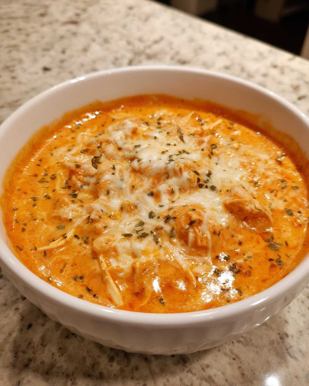 Close-up of a creamy, orange-colored buffalo chicken dip topped with melted white cheese and dried herbs in a white bowl.