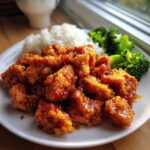 Close-up of crispy, glazed orange chicken recipe pieces served with white rice and steamed broccoli on a white plate.