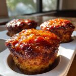 Three perfectly baked mini meatloaf portions in a white muffin tin, topped with a glistening, caramelized glaze.