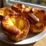 Five golden brown, perfectly risen yorkshire pudding cups sitting on a white plate near a window.