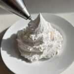 A piping bag tip is dispensing fluffy white vanilla buttercream frosting onto a white plate, creating a tall swirl.
