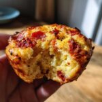 Close-up of a hand holding a savory pizza muffin with a bite taken out, showing a fluffy interior and pepperoni topping.