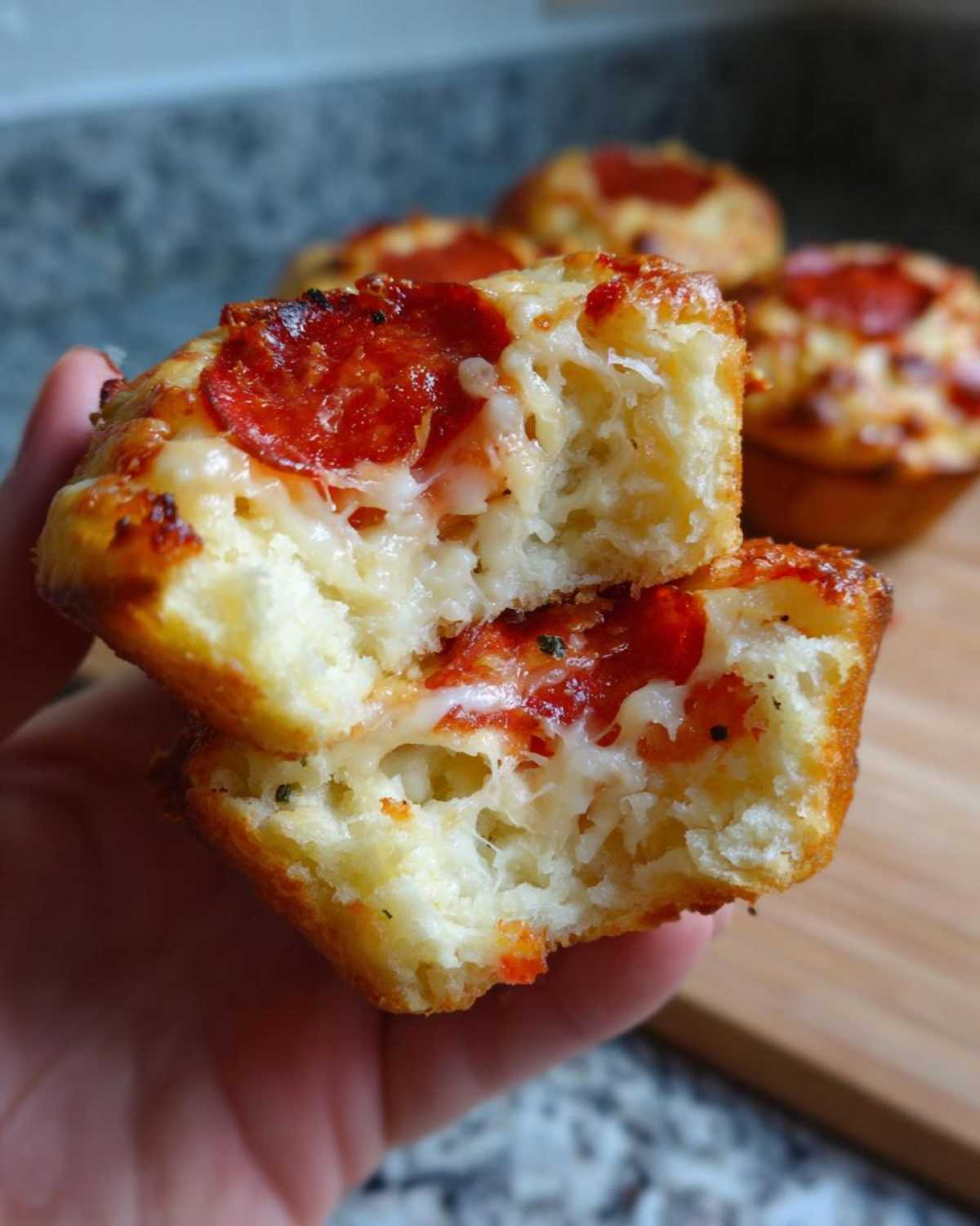 A hand holding a pizza muffin cut in half, showing the fluffy interior, melted cheese, and pepperoni topping of the pizza muffins.