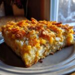 A thick slice of creamy potato casserole topped with golden, crunchy cornflakes and herbs, served on a plate.