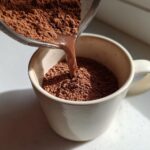 Close-up of rich brown hot cocoa mix being poured from a sieve into a white mug.