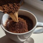 Close-up of homemade hot cocoa mix powder being poured into a white mug containing liquid.