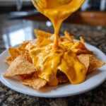 Thick, bright orange nacho cheese sauce being poured from a glass container onto a pile of tortilla chips on a white plate.