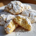 A stack of soft, yellow lemon crinkle cookies heavily dusted with powdered sugar, with one cookie broken open to show the texture.