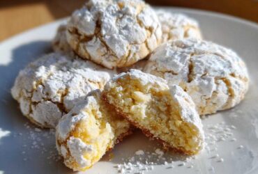 A stack of soft, yellow lemon crinkle cookies heavily dusted with powdered sugar, with one cookie broken open to show the texture.